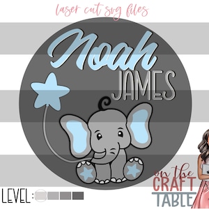 Könnte beinhalten: Ein grauer Kreis mit dem Text "Noah James" in weißer kursivschrift. Ein blauer Stern mit einer daran befestigten Schnur befindet sich auf der linken Seite des Kreises. Ein grauer Elefant mit blauen Ohren hält einen blauen Stern. Der Text "on the craft table" befindet sich unterhalb des Kreises.