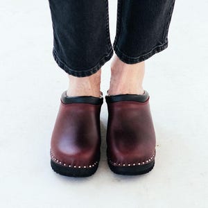 May include: A pair of burgundy leather clogs with black trim and a row of decorative studs around the sole.