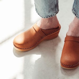 May include: A pair of brown leather clogs with a wooden sole and a strap across the foot. The clogs have small metal studs around the edge of the sole.