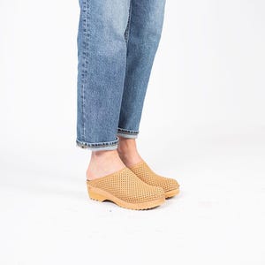 May include: Tan perforated clogs with a wooden sole. The clogs have a low heel and a slip-on design. The shoes are shown with blue jeans. The clogs are a neutral color and can be worn with many outfits.