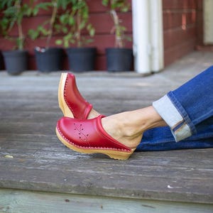 May include: Red clogs with a floral design on the toe, featuring a wooden sole and a strap. The clogs are paired with blue jeans. The clogs are a classic style of footwear.