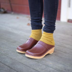 Pode incluir: Sapatos de couro marrom com solas de madeira e um pequeno salto. Os sapatos são usados com meias amarelo mostarda e calças de ganga azul escuro.