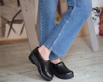 May include: Black leather clogs with a rounded toe and a small heel. The clogs have a decorative row of studs around the edge of the sole.