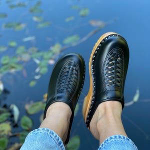 Puede incluir: Un par de zuecos de cuero negro con un diseño tejido en la parte superior. Los zuecos tienen una base de madera y los lleva una persona con pantalones vaqueros azules.