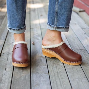 May include: Brown leather clogs with a shearling lining and wooden soles. The clogs have a strap across the top and are studded around the sole. The person is wearing blue jeans.