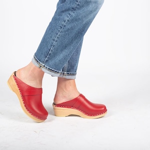 Peut inclure: Une paire de sabots en cuir rouge avec des semelles en bois. Les sabots ont une sangle sur le dessus du pied et sont portés par une personne en jean bleu.