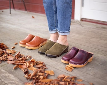 May include: Three pairs of wooden clogs, one pair is brown leather, one pair is olive green suede, and one pair is burgundy leather.