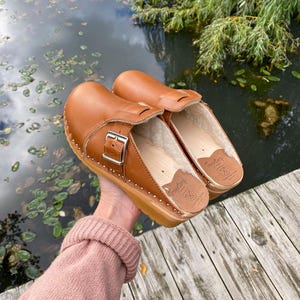 Pode incluir: Um par de tamancos de couro castanho com tachas e fivelas prateadas. Os tamancos têm sola de madeira e interior macio de cor clara. Os tamancos são mostrados ao ar livre, perto de um corpo de água.