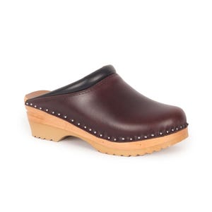 Puede incluir: Zueco de cuero burdeos con cuello negro y suela de madera. El zapato presenta tachuelas plateadas alrededor del borde de la parte superior y un tacón de madera marrón claro. El zueco está diseñado para la comodidad y el estilo.