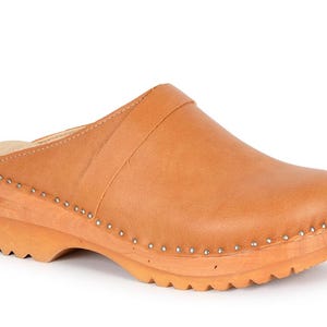 Könnte beinhalten: Ein hellbrauner Lederclog mit Holzsohle. Der Schuh hat eine runde Spitze, einen Riemen über dem Oberteil und ist mit kleinen silbernen Nieten befestigt. Die Sohle ist aus hellem Holz und hat einen geriffelten Boden.