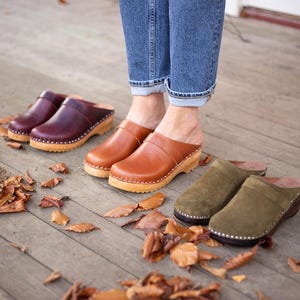 May include: Four pairs of leather clogs in brown, burgundy, and olive green. The clogs have wooden soles and are worn by a person standing on a wooden floor.