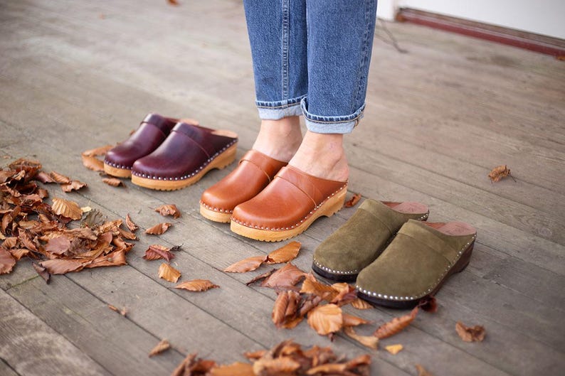 May include: Four pairs of leather clogs in brown, burgundy, and olive green. The clogs have wooden soles and are worn by a person standing on a wooden floor.