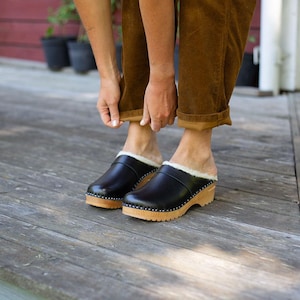 Pode incluir: Tamancos de couro preto com sola de madeira e forro de pele de carneiro. Os tamancos têm bico arredondado e são presos com pequenos pregos prateados ao redor da sola. O salto tem cerca de 5 cm.