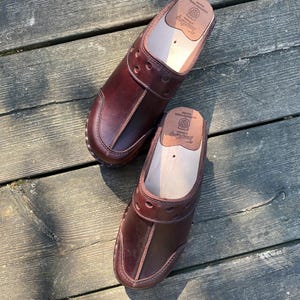 May include: A pair of brown leather clogs with a wooden sole. The clogs have a strap across the top and a decorative pattern of holes. The clogs are on a wooden surface.