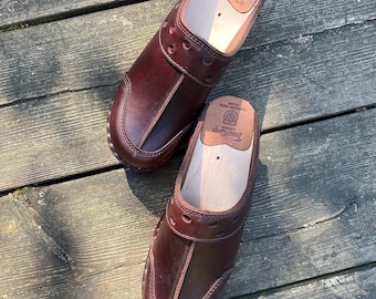 May include: A pair of brown leather clogs with a wooden sole. The clogs have a strap across the top and a decorative pattern of holes. The clogs are on a wooden surface.