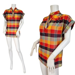 May include: A short-sleeved plaid shirt in a vibrant mix of colors, including red, yellow, orange, and green. The shirt features a mandarin collar and a buttoned placket. The fabric has a woven texture, and the shirt is displayed on a mannequin.