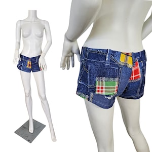 May include: Blue denim shorts with patchwork details. The shorts feature colorful plaid patches in red, yellow, and green. The shorts are displayed on a mannequin.