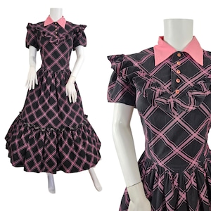 May include: A black dress featuring a pink plaid pattern, a solid pink collar, and short puff sleeves. The bodice includes a button-up placket with pink buttons and ruffled trim across the chest. The skirt is full with a ruffled hemline.