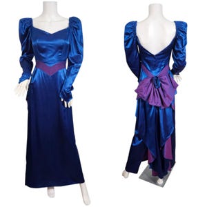 May include: A long, royal blue satin gown with long sleeves and a V-neckline. The dress features a purple accent at the waist and a large bow detail on the back. The dress is displayed on a mannequin.