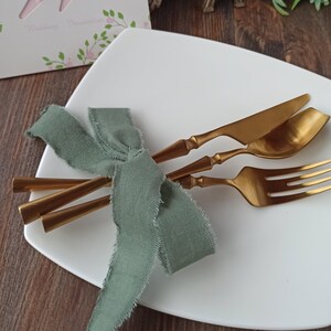 May include: A white square plate with a gold knife, fork, and spoon tied together with a sage green ribbon. The plate is on a wooden surface.