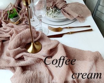 May include: A table setting with a coffee cream colored gauze table runner. Gold-colored flatware, candle holders, and a geometric terrarium are arranged on a white tablecloth. The text "Coffee cream" is visible on the table runner.