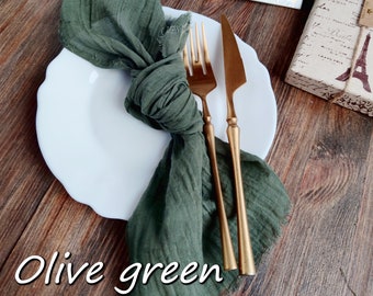 May include: Olive green fabric napkin folded and placed on a white plate with gold cutlery. The napkin is on a wooden table.