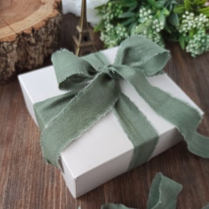 May include: A white gift box tied with a sage green ribbon. The ribbon is tied in a bow and the ends are frayed.