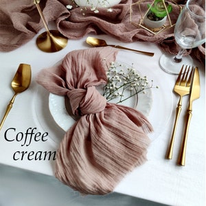 May include: A table setting with a coffee cream colored runner and napkin. Gold-colored flatware, a candle holder, and a geometric terrarium are arranged on a white tablecloth. A white vase holds white flowers.