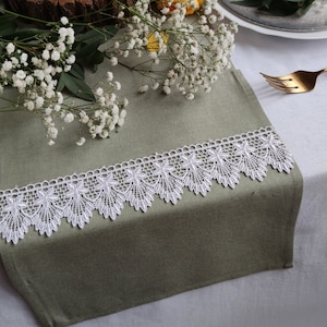 May include: A green linen table runner with white lace trim. The runner is on a white tablecloth with a wooden slice, a candle, and white flowers.