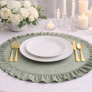 May include: A table setting featuring a sage green ruffled placemat, white plates, and gold cutlery. A bouquet of white flowers and lit candles complete the elegant dining scene. The placemat is approximately 15 inches in diameter.