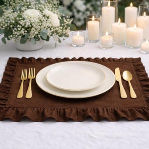 Peut inclure: Une table dressée avec un set de table marron à volants, des assiettes blanches à bord doré et des couverts dorés. Des bougies allumées dans des supports en verre et une composition florale blanche créent une ambiance élégante.