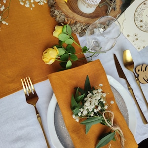 May include: A mustard yellow linen napkin with a sprig of white flowers and green leaves tied with twine, placed on a white plate with a gray rim. The plate is on a white tablecloth with a gold fork and knife.