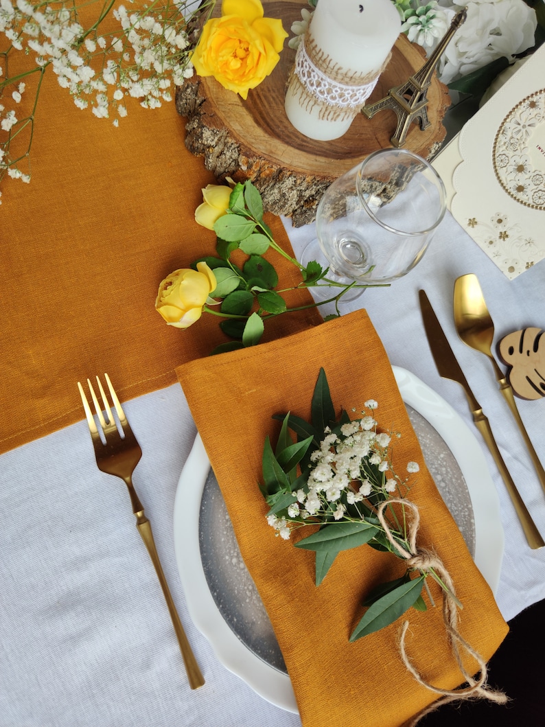 May include: A mustard yellow linen napkin with a sprig of white flowers and green leaves tied with twine, placed on a white plate with a gray rim. The plate is on a white tablecloth with a gold fork and knife.