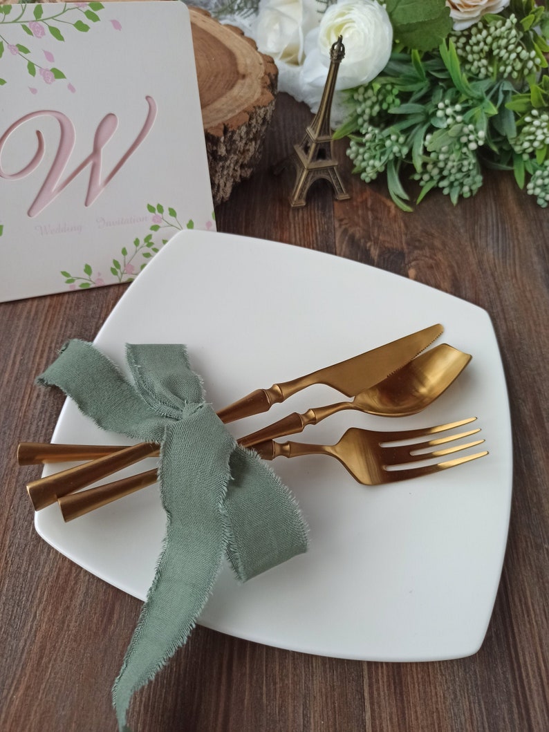May include: A white square plate with a gold knife, fork, and spoon tied together with a sage green ribbon. The plate is on a wooden surface.