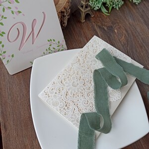 May include: A white wedding invitation with a lace design and a green ribbon. The invitation is on a white plate with a wooden background.