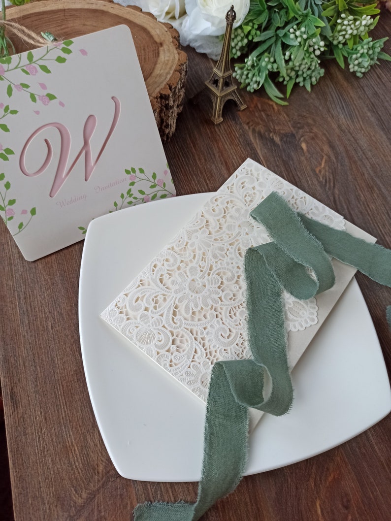 May include: A white wedding invitation with a lace design and a green ribbon. The invitation is on a white plate with a wooden background.