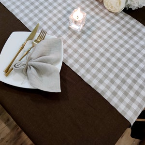 May include: A brown linen tablecloth with a white and gray gingham table runner. A white plate with gold silverware and a linen napkin is on the table runner. A small glass candle holder with a lit candle is on the table runner.