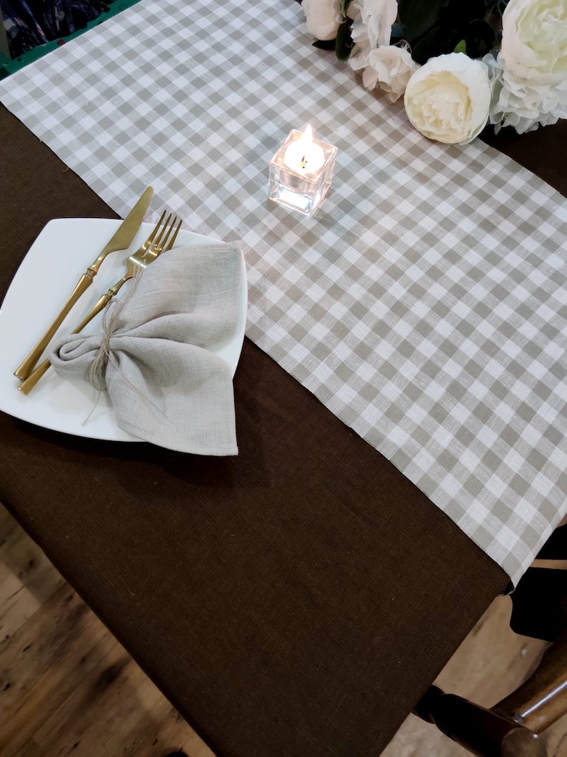 May include: A brown linen tablecloth with a white and gray gingham table runner. A white plate with gold silverware and a linen napkin is on the table runner. A small glass candle holder with a lit candle is on the table runner.