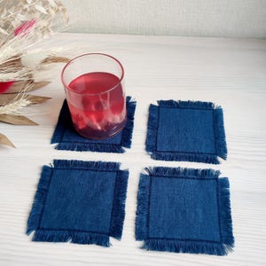 Puede incluir: Cuatro posavasos de lino azul con bordes deshilachados. Los posavasos están dispuestos en forma de cuadrado sobre una superficie de madera blanca. Un vaso de líquido rojo se encuentra sobre uno de los posavasos.