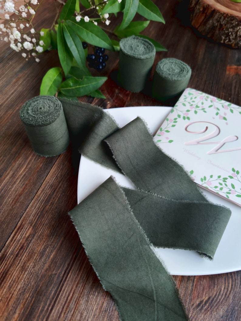 May include: A green silk ribbon with frayed edges on a white plate. The ribbon is rolled up in the background. The plate is on a wooden surface.