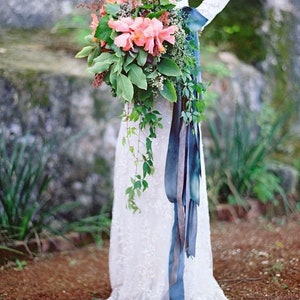 May include: A bride holds a cascading bouquet of pink and orange flowers with greenery and a long blue ribbon.