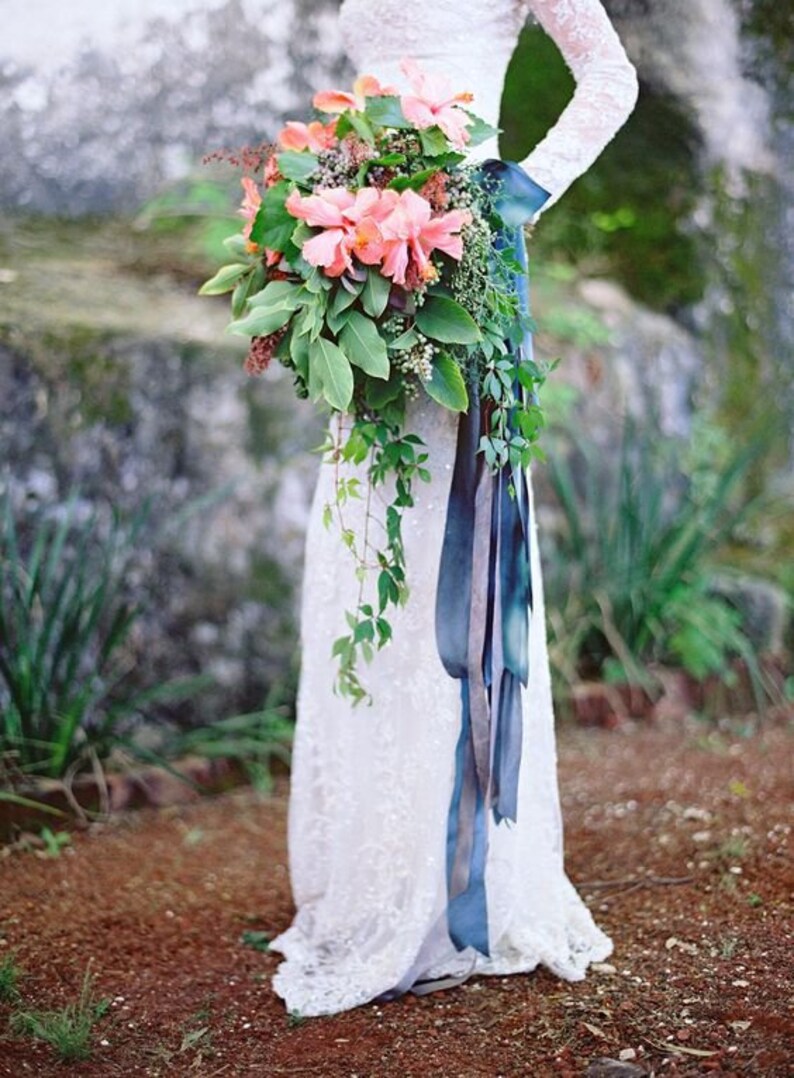 May include: A bride holds a cascading bouquet of pink and orange flowers with greenery and a long blue ribbon.