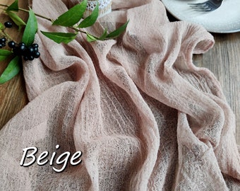 May include: Beige table runner made of a sheer, gauzy fabric with a delicate texture. The runner is draped over a wooden table and has a candle in the center.