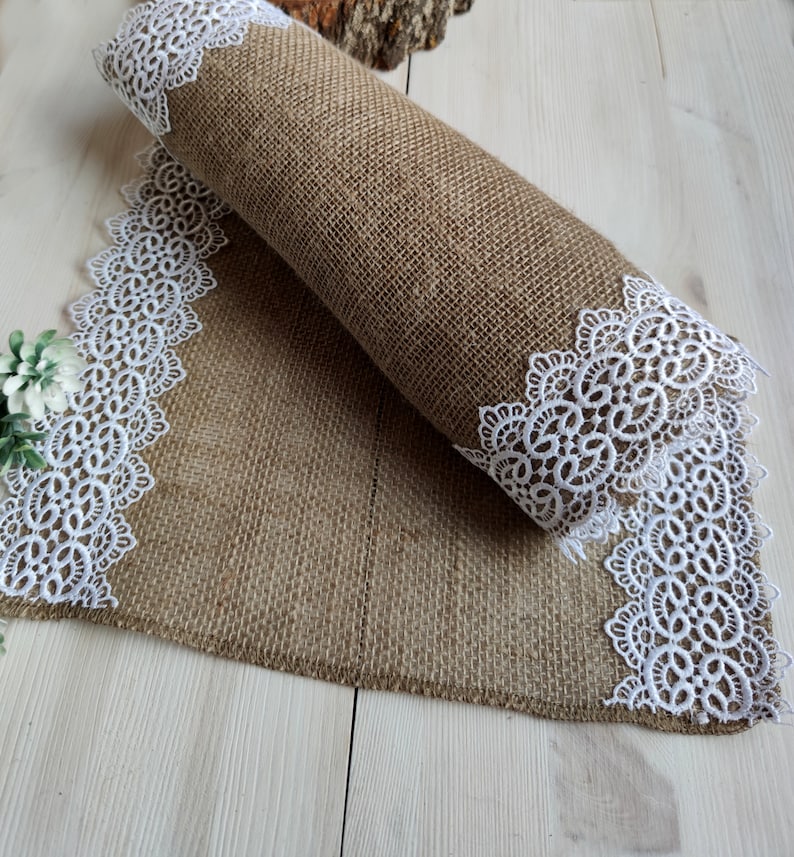 Peut inclure: Un chemin de table en toile de jute brune avec une bordure en dentelle blanche. Le chemin de table est roul&eacute; et la bordure en dentelle est visible aux deux extr&eacute;mit&eacute;s.