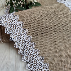Peut inclure: Un chemin de table en toile de jute avec une bordure en dentelle blanche. Le chemin de table est pli&eacute; en deux et la bordure en dentelle est visible des deux c&ocirc;t&eacute;s.