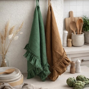 a kitchen counter with two green and brown towels hanging on a rack, along with various kitchen utensils and decor items such as a vase with dried flowers, a wooden cutting board, and a bowl of fresh artichokes.