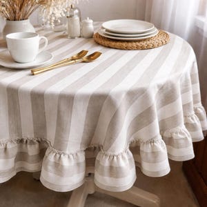 a table setting with a white tablecloth, a white plate, a white cup, and a set of gold-colored spoons. The table is set with a woven placemat and a white vase with dried flowers.