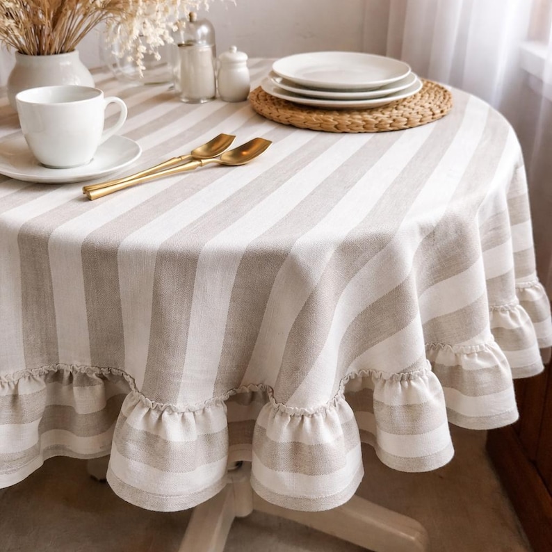 a table setting with a white tablecloth, a white plate, a white cup, and a set of gold-colored spoons. The table is set with a woven placemat and a white vase with dried flowers.