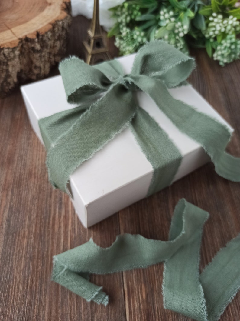 May include: A white gift box tied with a sage green ribbon. The ribbon is tied in a bow and the ends are frayed.