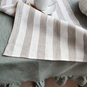 May include: A white and beige striped linen table runner with a ruffled green tablecloth underneath. A white coffee cup and saucer are on top of the table runner.